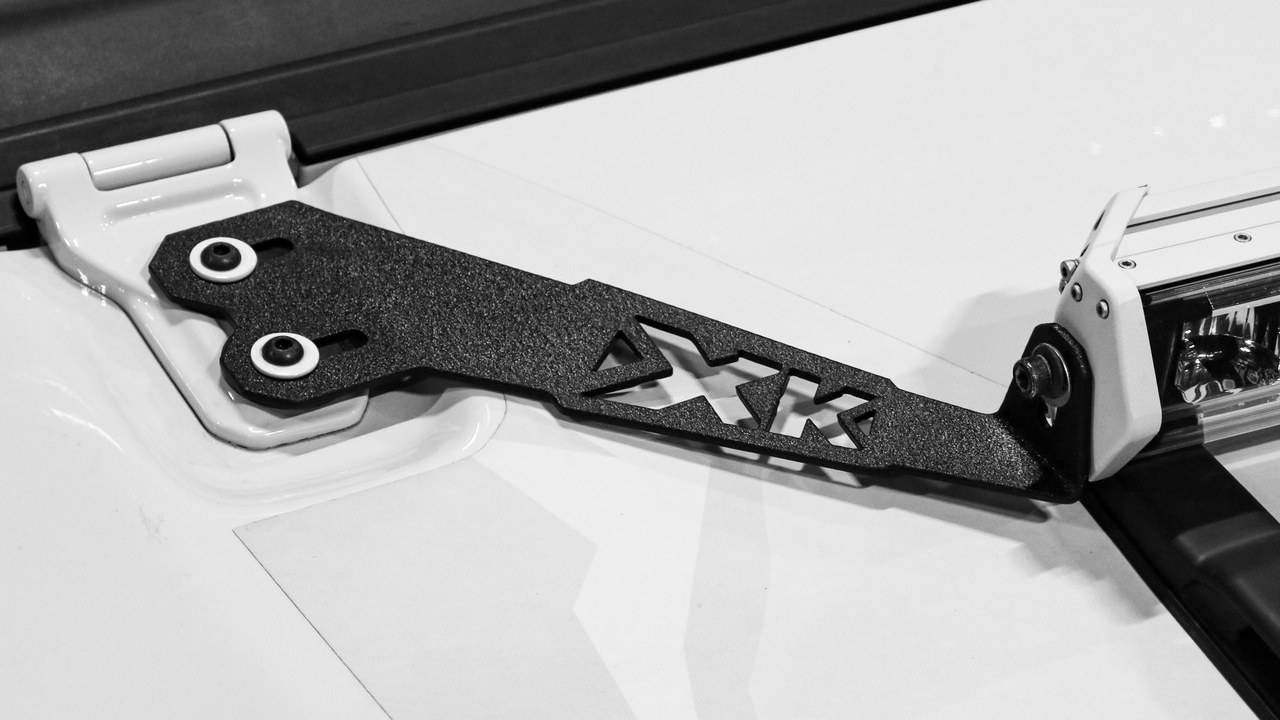 Black steel bracket stamped XK, bolted to a white hood hinge and holding the end of a light bar